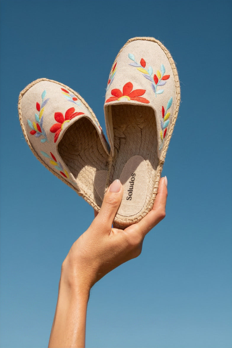 A hand holds a pair of Soludos espadrille slip-on shoes against a clear blue sky. The shoes have a beige woven jute upper with colorful embroidered floral patterns in red, orange, yellow, and blue.