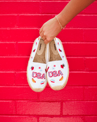 White espadrilles with colorful USA embroidery on them being held in front of a red wall